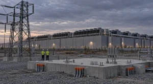 Two utility workers stand beside an empty concrete transformer pad with capped conduit stubs, while a finished hyperscale data center waits under an overcast dusk sky.