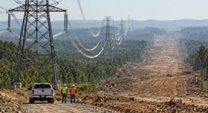 Heat-sagged conductors hang from lattice towers across forested hills beside a freshly cleared, still-empty parallel right-of-way, with two engineers and a pickup truck in the foreground.