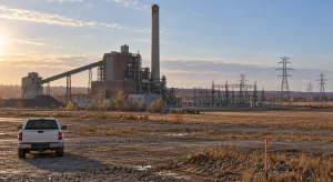 A coal power station and electrical substation stand beyond surveyed rural ground, with a white pickup truck in the foreground under warm evening light.
